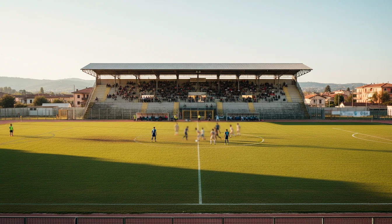 Stadio di calcio di provincia con tribuna coperta durante una partita diurna